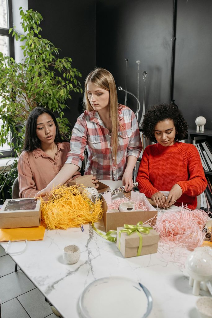 Three women of diverse backgrounds wrapping gifts together in a cozy indoor setting.